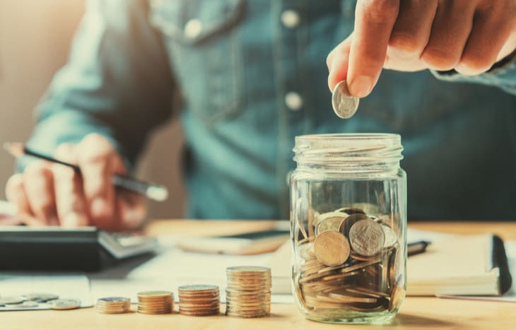 A person putting money in a jar on top of coins.